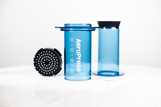Cafetera manual AeroPress Clear en color azul translúcido. Despiece del método de preparación que incluye cámara, émbolo y tapa de filtro para café de especialidad.